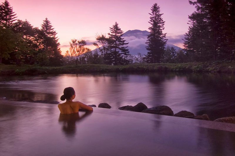 A woman in the female onsen at Hilton Niseko Village at sunset with a view of Mt Yotei and surrounded by forest.